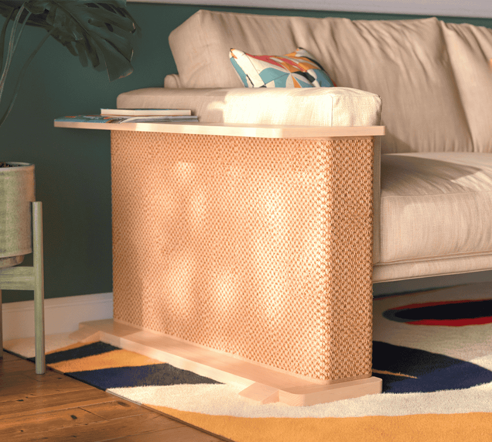 A Cat-e-Corner table placed next to a sofa, integrating a cat's scratching post with a small table, in a room with a plant and a colorful cushion.